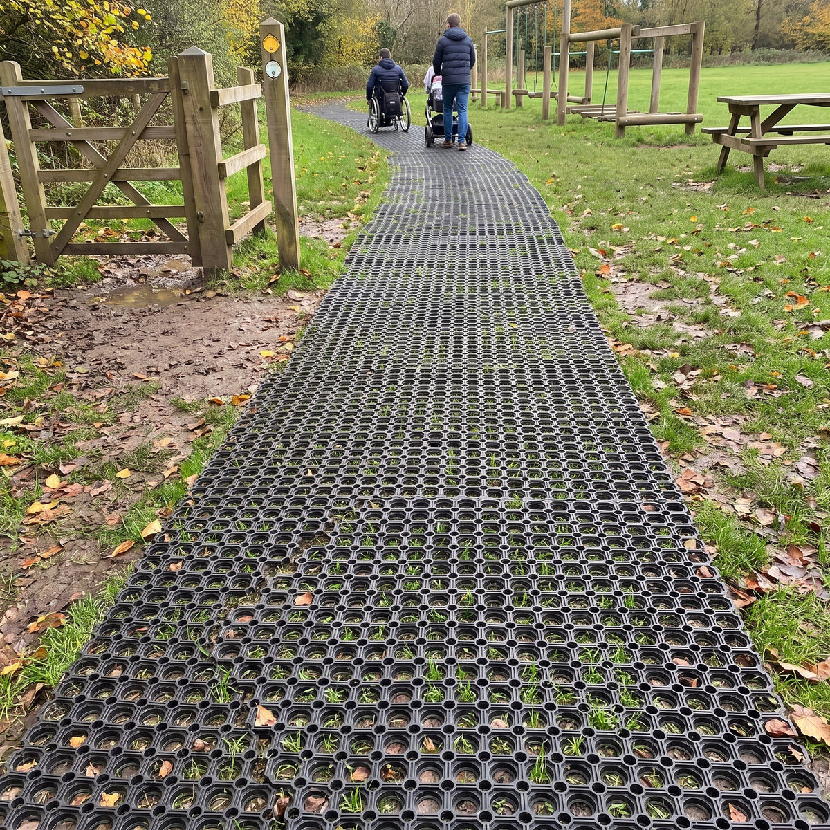 Wheelchair-accessible path of rubber grass mats across a muddy park field, with a wheelchair user and a pushchair on the firm mat surface
