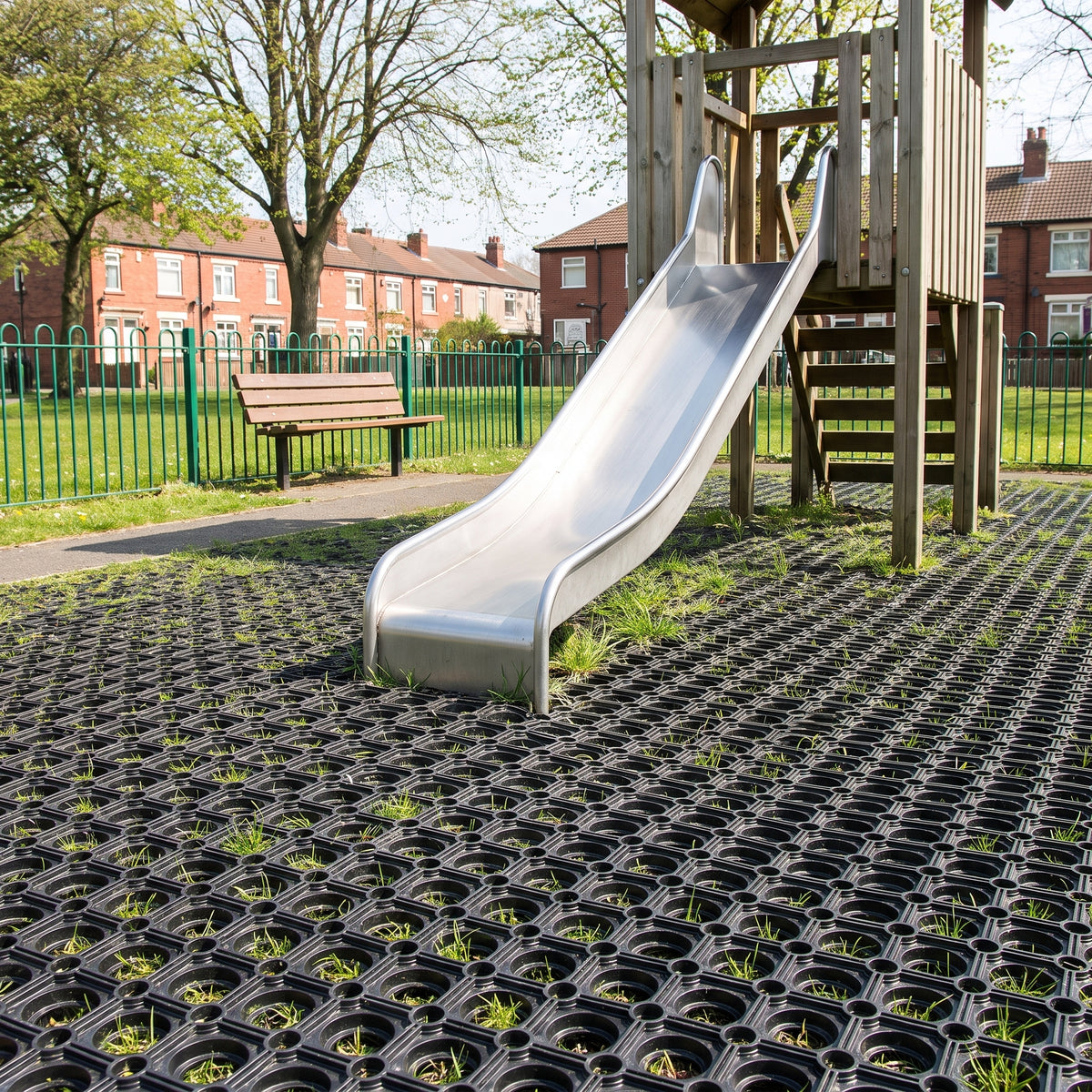 Black rubber grass mats laid as safety surfacing under a timber play tower and stainless steel slide in a fenced council park playground, with grass growing up through the mats