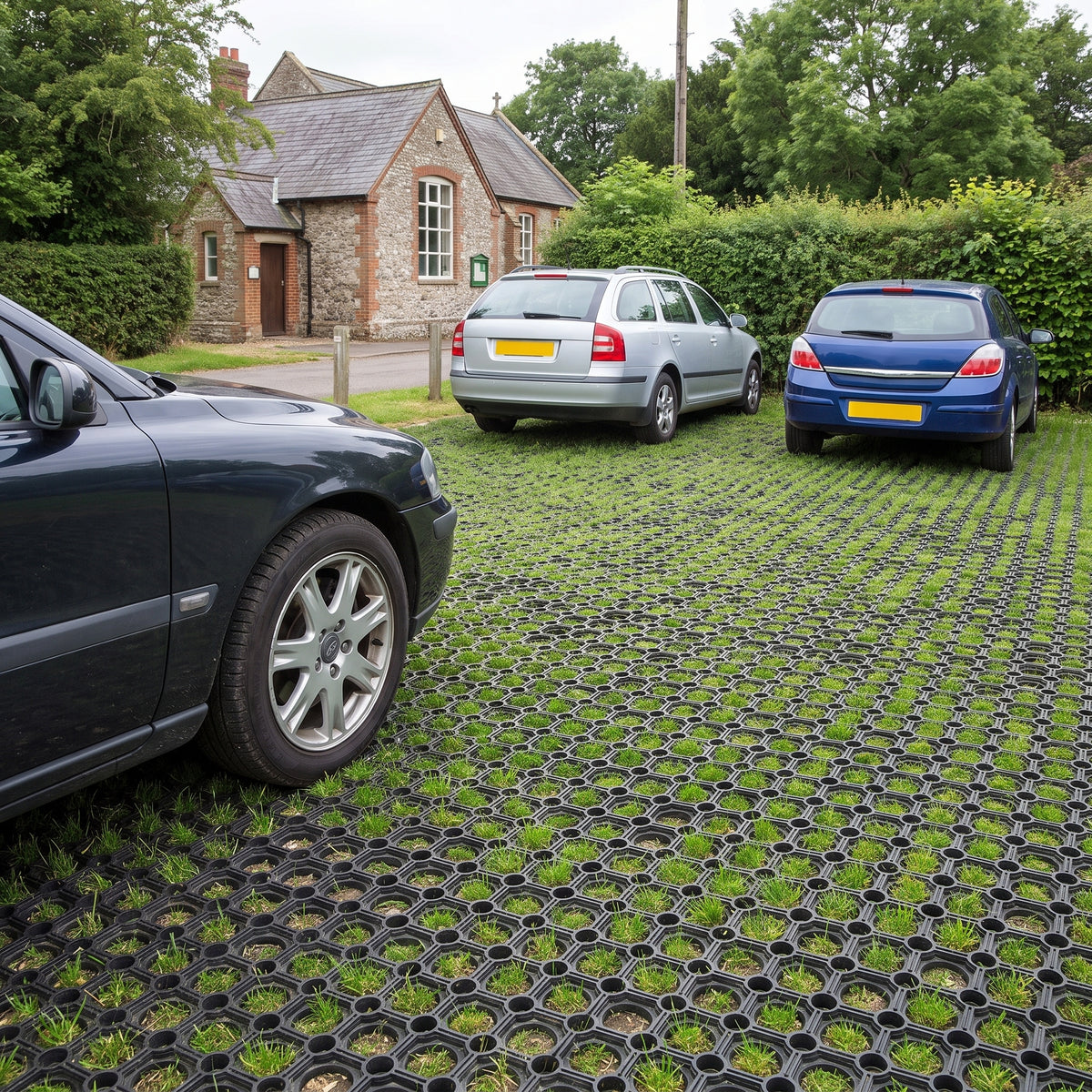 Cars parked on rubber grass mats in a grass car park beside a village hall, with grass growing through the mats
